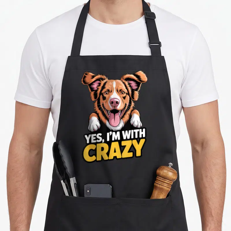 Dog cotton duck aprons - Cartoon dog peeking over Yes I'm With Crazy text — shown on a black cotton canvas apron against a white background