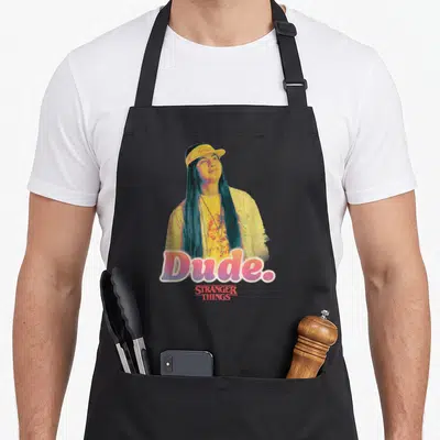 Argyle dude apron - Portrait of man with long hair yellow cap and Dude Stranger Things text — shown on a black cotton canvas apron against a white background