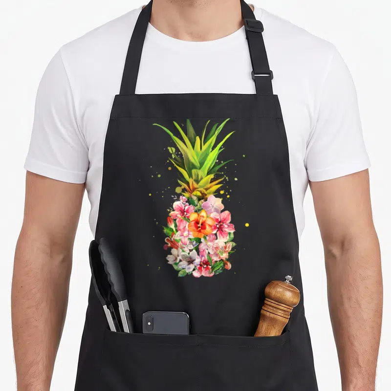 Flower pineapple apron - Watercolor pineapple made of pink and orange flowers with green leaves and scattered yellow — shown on a black cotton canvas apron against a white background