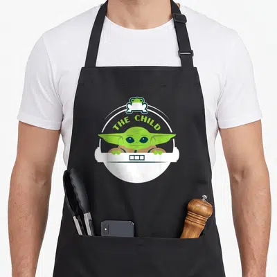 Pod frog aprons - Cartoon alien child in white pod with THE CHILD text and small frog on top border — shown on a black cotton canvas apron against a white background