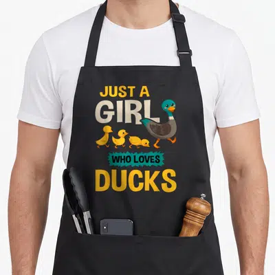 Retro duck apron - Just A Girl Who Loves Ducks text with cartoon duck and three yellow ducklings walking — shown on a black cotton canvas apron against a white background