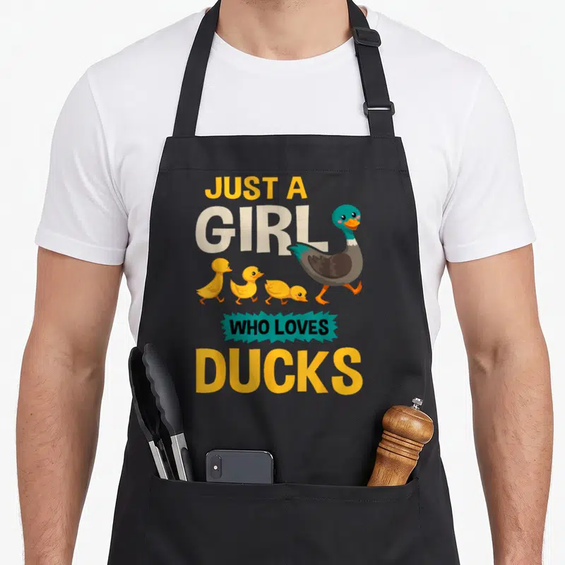 Retro duck apron - Just A Girl Who Loves Ducks text with cartoon duck and three yellow ducklings walking — shown on a black cotton canvas apron against a white background