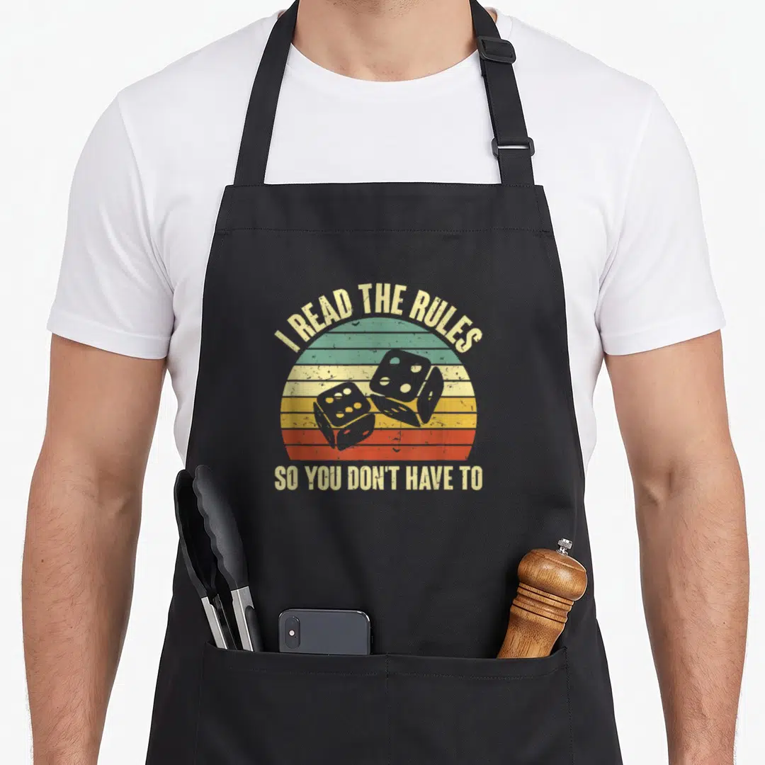 Night game aprons - Two grey dice on a striped beige green yellow orange red sunset with arched text above and straight — shown on a black cotton canvas apron against a white background
