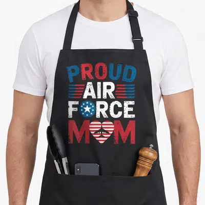 Flag air force mom apron - PROUD AIR FORCE MOM text in red white and blue with star circle and airplane heart replacing — shown on a black cotton canvas apron against a white background