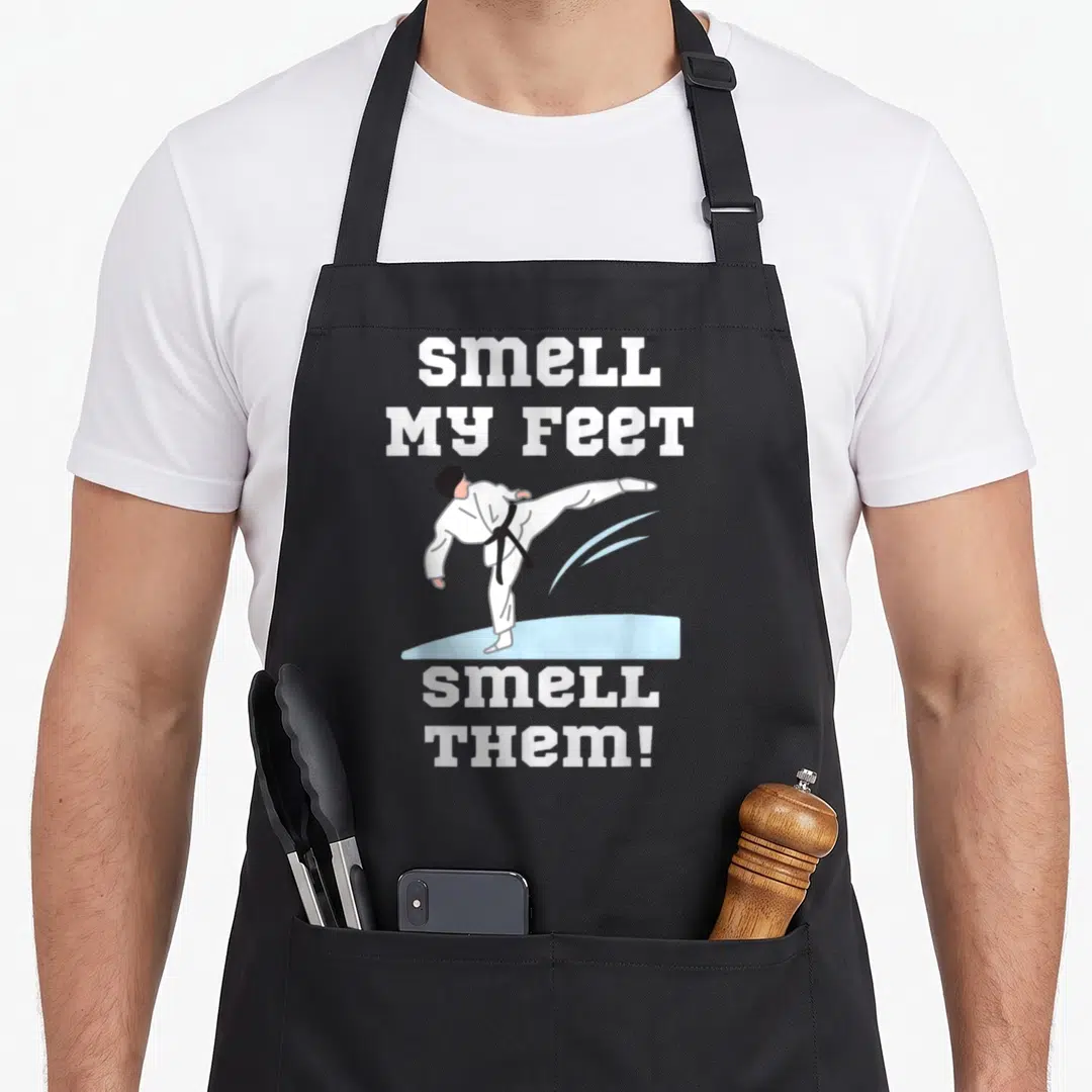 Smell infantry apron - Cartoon karateka high kick with SMELL MY FEET SMELL THEM text in white, black, and light blue — shown on a black cotton canvas apron against a white background