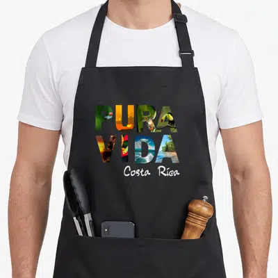 Pura friends apron - Pura Vida letters filled with toucan frog sunset beach photos above Costa Rica script — shown on a black cotton canvas apron against a white background