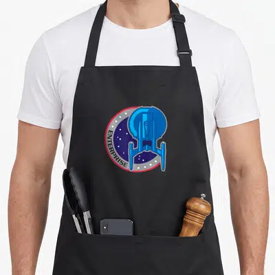 Funny enterprise star apron - Blue spaceship over dark blue circle with white stars and curved ENTERPRISE text bordered — shown on a black cotton canvas apron against a white background