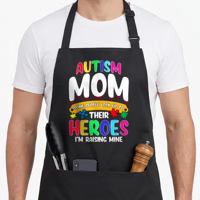 Their autism apron - Stacked text Autism Mom Heroes I'm Raising Mine with four colorful puzzle pieces — shown on a black cotton canvas apron against a white background