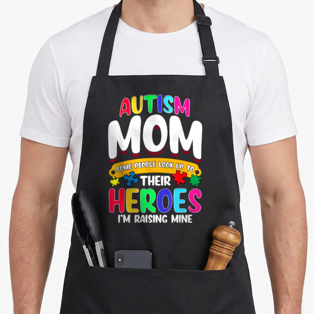 Their autism apron - Stacked text Autism Mom Heroes I'm Raising Mine with four colorful puzzle pieces — shown on a black cotton canvas apron against a white background