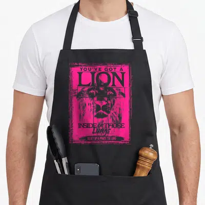 Those lion apron - Pink distressed lion head with stacked text You've Got A Lion Inside Of Those Lungs So Get Up — shown on a black cotton canvas apron against a white background