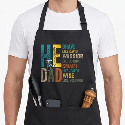 Christian smart apron - He Is Dad Brave Like David Warrior Like Joshua Smart Like Joseph Wise Like Solomon distressed — shown on a black cotton canvas apron against a white background