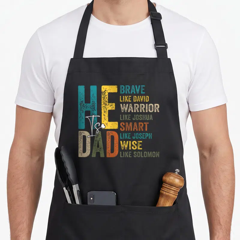 Christian smart apron - He Is Dad Brave Like David Warrior Like Joshua Smart Like Joseph Wise Like Solomon distressed — shown on a black cotton canvas apron against a white background