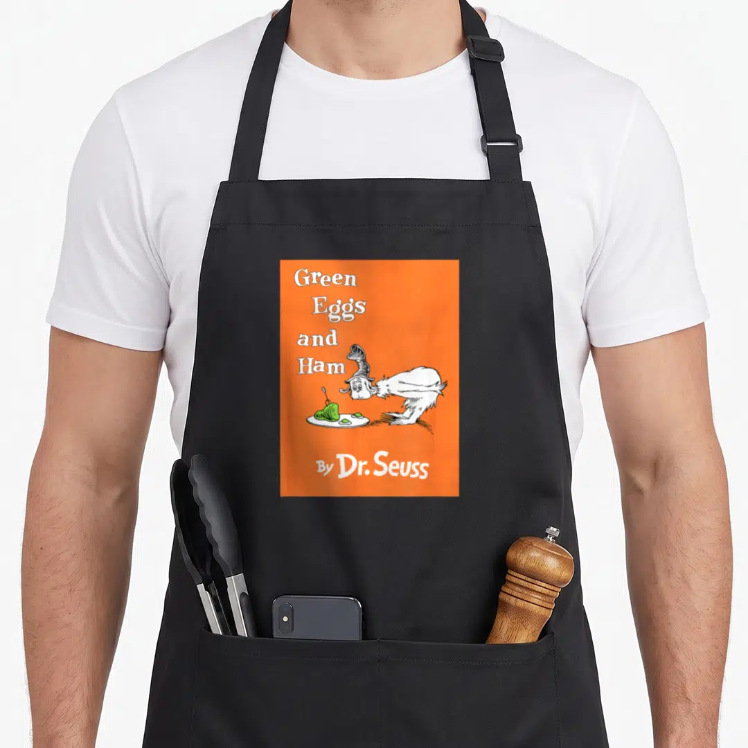 Book egg apron - Orange background with white creature and Green Eggs and Ham title text — shown on a black cotton canvas apron against a white background