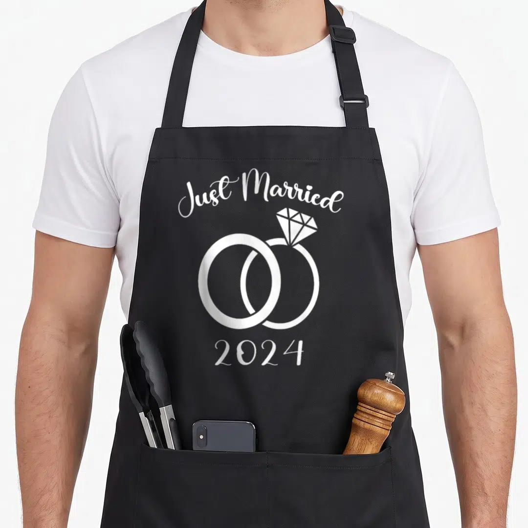 Ring newlywed aprons - Just Married 2024 text above two interlocking white rings with diamond accent — shown on a black cotton canvas apron against a white background