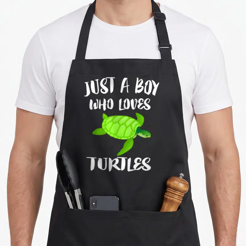 Sea animal apron - Green cartoon sea turtle centered between white text Just A Boy Who Loves Turtles — shown on a black cotton canvas apron against a white background
