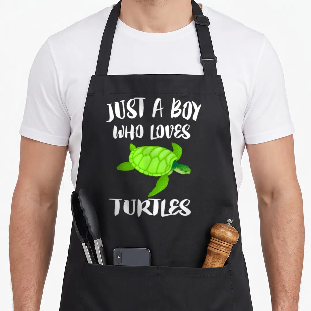 Sea animal apron - Green cartoon sea turtle centered between white text Just A Boy Who Loves Turtles — shown on a black cotton canvas apron against a white background