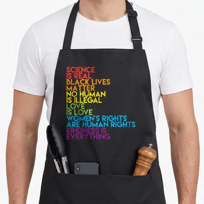 Matter science apron - Rainbow stacked text: Science Is Real, Black Lives Matter, No Human Is Illegal, Love Is Love — shown on a black cotton canvas apron against a white background