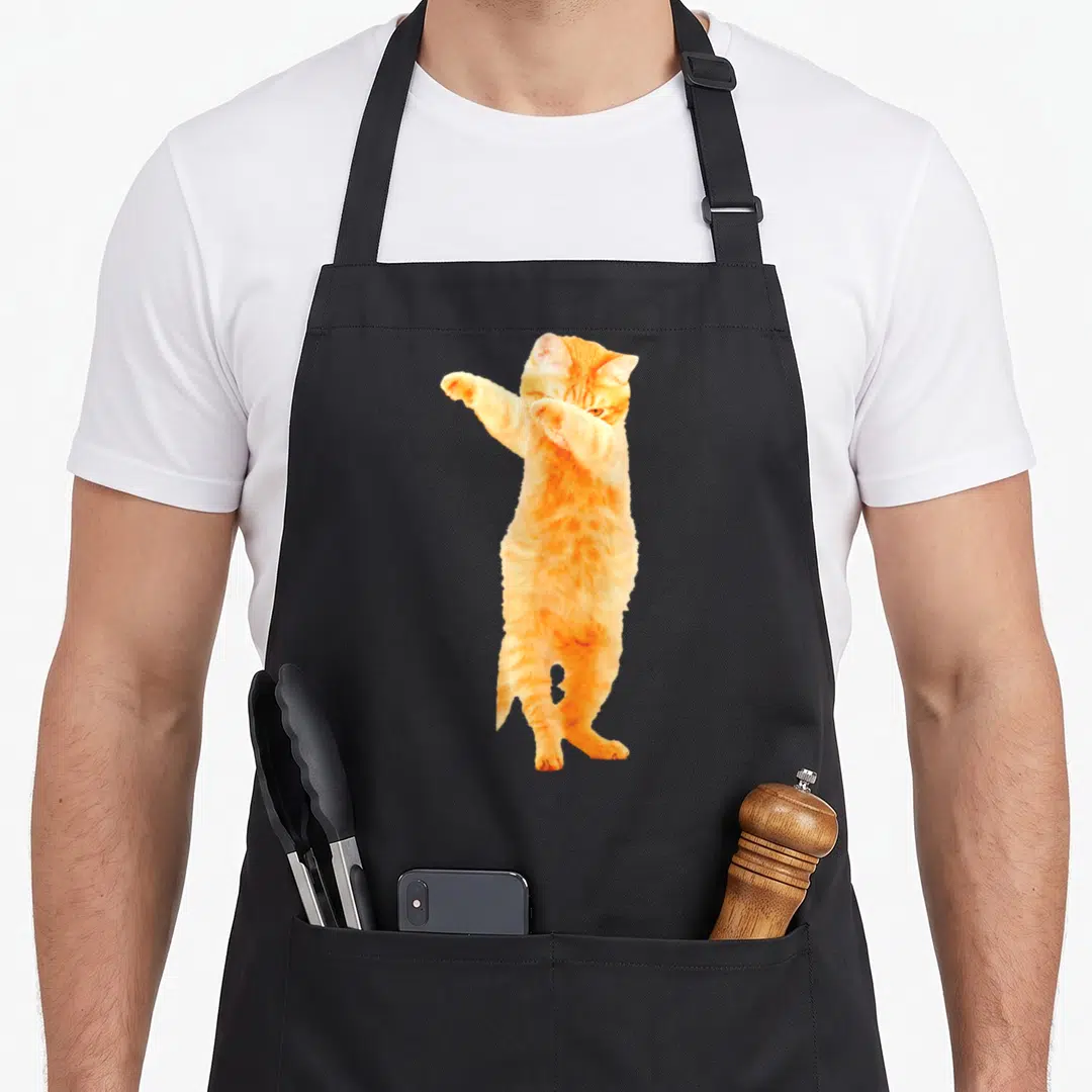 Dabbing kitty cat apron - Orange cat standing on hind legs with one paw raised in a dab pose — shown on a black cotton canvas apron against a white background