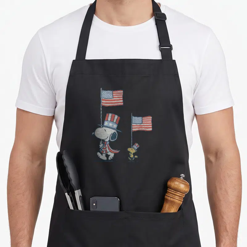 Woodstock decade apron - Snoopy and Woodstock in patriotic top hats holding American flags in red white blue and yellow — shown on a black cotton canvas apron against a white background