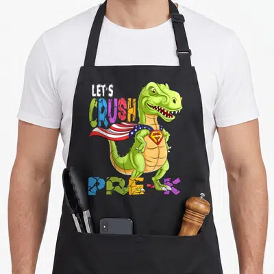 Ready crush preschool apron - Cartoon green T-Rex in cape with cracked multicolor text LET'S CRUSH PRE-K — shown on a black cotton canvas apron against a white background