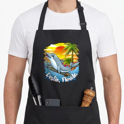 Destin spring apron - Cartoon dolphin surfing on a wave below curved Destin Florida text and palm trees — shown on a black cotton canvas apron against a white background
