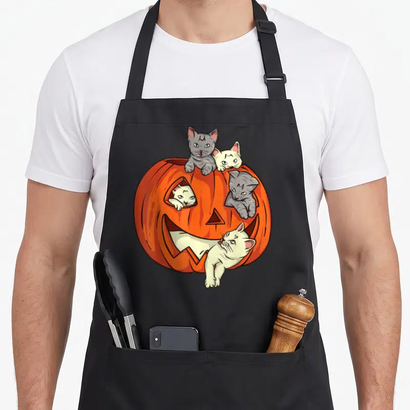 Pumpkin cat apron - Four cartoon cats peeking from orange jack o lantern holes on black apron — shown on a black cotton canvas apron against a white background
