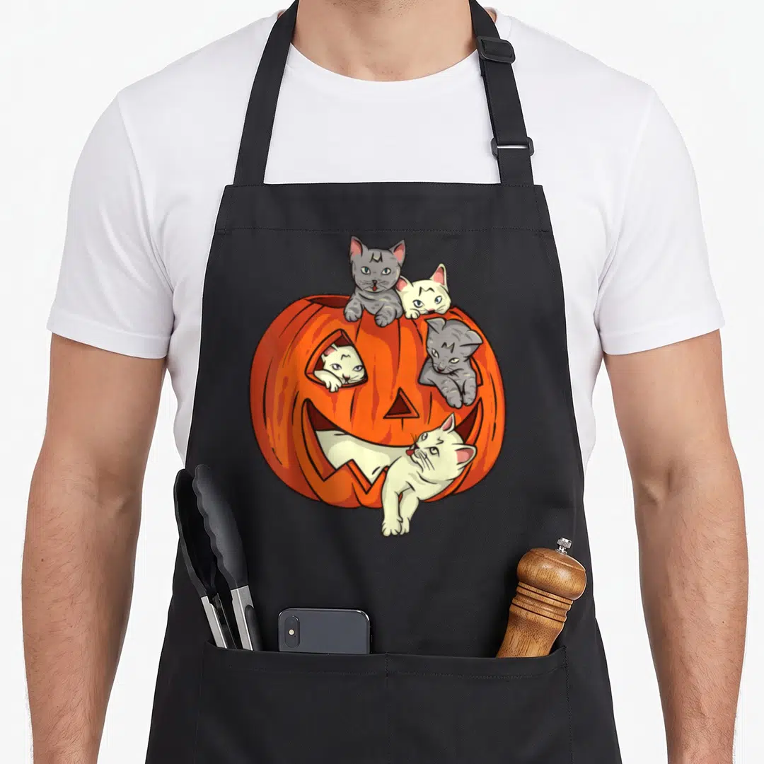 Pumpkin cat apron - Four cartoon cats peeking from orange jack o lantern holes on black apron — shown on a black cotton canvas apron against a white background