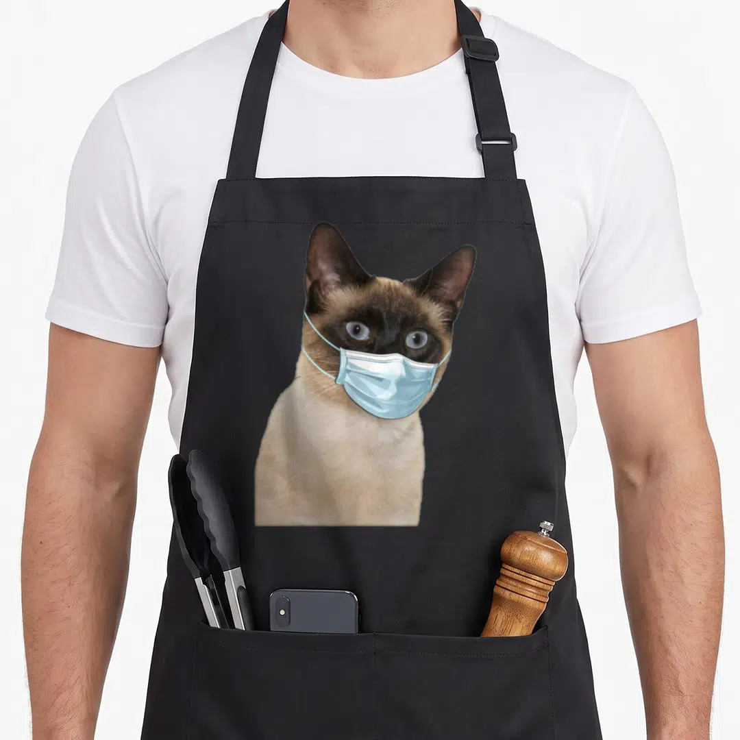 Face cute cat aprons - Realistic Siamese cat wearing light blue surgical mask over muzzle — shown on a black cotton canvas apron against a white background