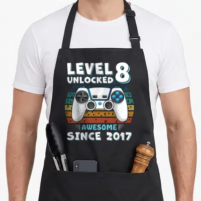 8yr birthday apron - White game controller over blue orange pixel blocks with Level 8 Unlocked Since 2017 text — shown on a black cotton canvas apron against a white background