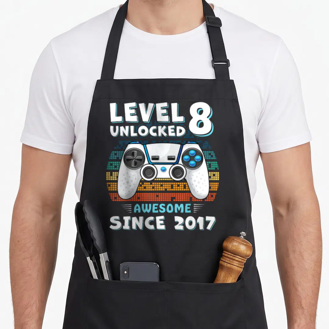 8yr birthday apron - White game controller over blue orange pixel blocks with Level 8 Unlocked Since 2017 text — shown on a black cotton canvas apron against a white background
