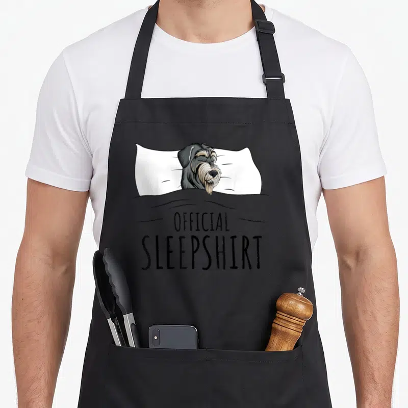 Sleepshirt miniature schnauzer apron - Line art of dog on pillow under grey blanket with official sleepshirt text — shown on a black cotton canvas apron against a white background
