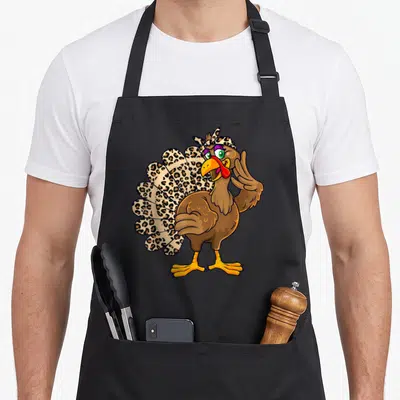 Thanksgiving fall aprons - Cartoon turkey standing with leopard print tail and headscarf raising one wing — shown on a black cotton canvas apron against a white background