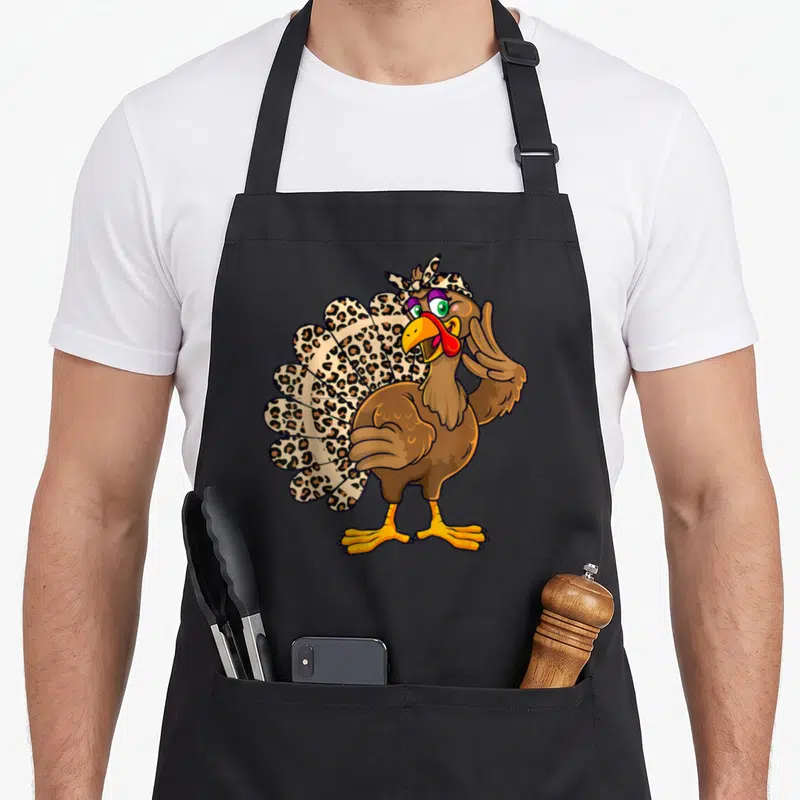 Thanksgiving fall aprons - Cartoon turkey standing with leopard print tail and headscarf raising one wing — shown on a black cotton canvas apron against a white background