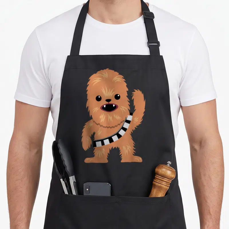 Cutie cartoon apron - Cartoon Chewbacca standing facing forward with striped bandolier in orange-brown and black — shown on a black cotton canvas apron against a white background