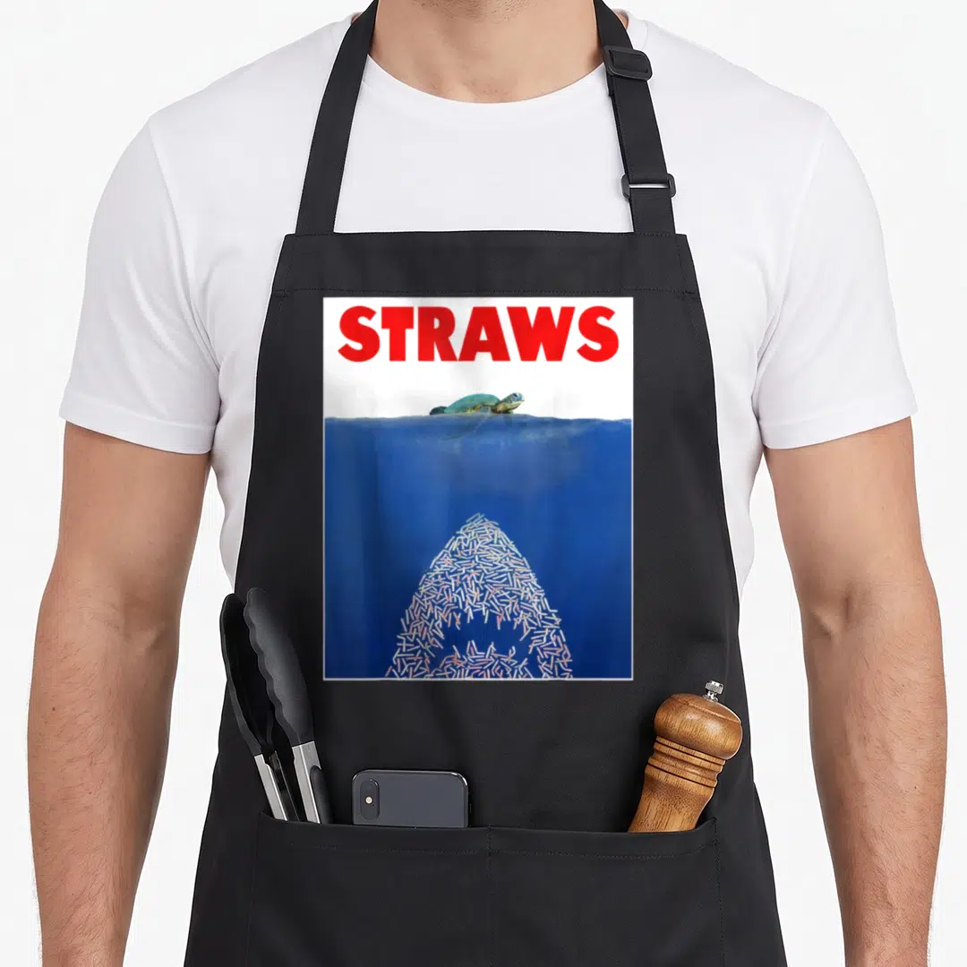 Save conservation apron - Sea turtle swims above a shark shape formed by straws under blue water with red text — shown on a black cotton canvas apron against a white background