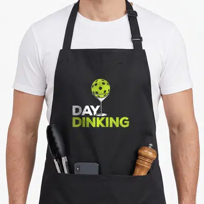 Dinking pickle apron - Lime green disc golf ball in white martini glass with distressed DAY DINKING text — shown on a black cotton canvas apron against a white background