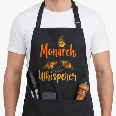 Entomologist butterfly apron - Three monarch butterflies surrounding stacked Monarch Whisperer text in orange and yellow — shown on a black cotton canvas apron against a white background