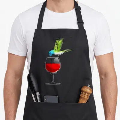 Funny hummingbird lover apron - Realistic hummingbird with green wings perched on wine glass rim dipping beak into red — shown on a black cotton canvas apron against a white background