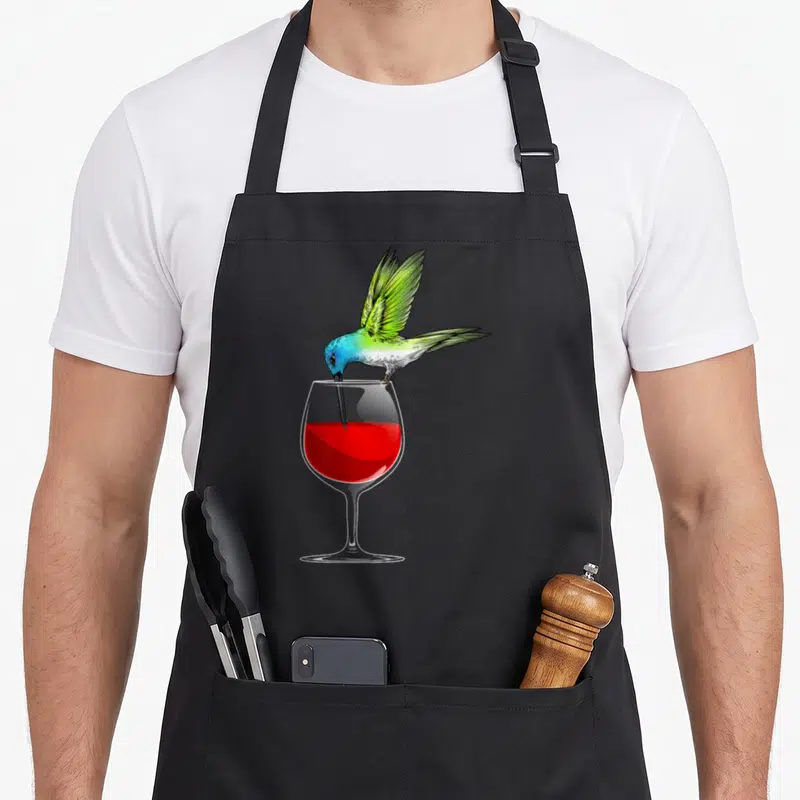 Funny hummingbird lover apron - Realistic hummingbird with green wings perched on wine glass rim dipping beak into red — shown on a black cotton canvas apron against a white background