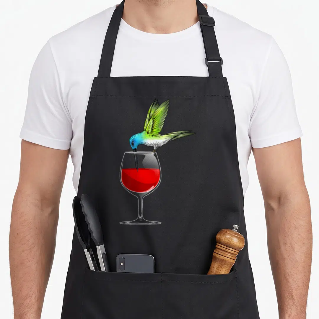 Funny hummingbird lover apron - Realistic hummingbird with green wings perched on wine glass rim dipping beak into red — shown on a black cotton canvas apron against a white background