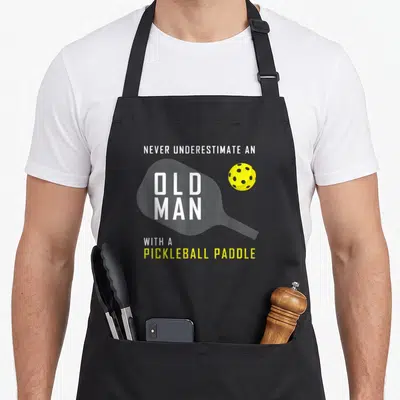 Underestimate pickleball apron - Stacked text reading Never Underestimate An Old Man With A Pickleball Paddle — shown on a black cotton canvas apron against a white background