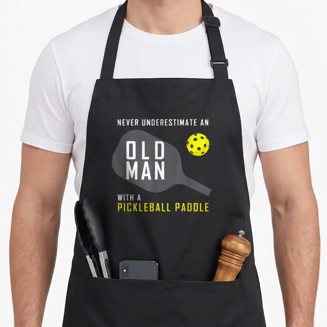 Underestimate pickleball apron - Stacked text reading Never Underestimate An Old Man With A Pickleball Paddle — shown on a black cotton canvas apron against a white background