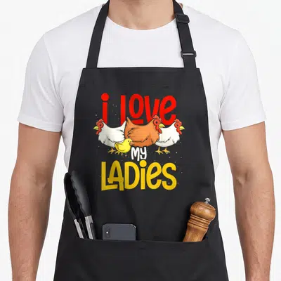 Farmer animal chicken apron - Three cartoon chickens and a chick surrounded by i love my ladies text in red and yellow — shown on a black cotton canvas apron against a white background