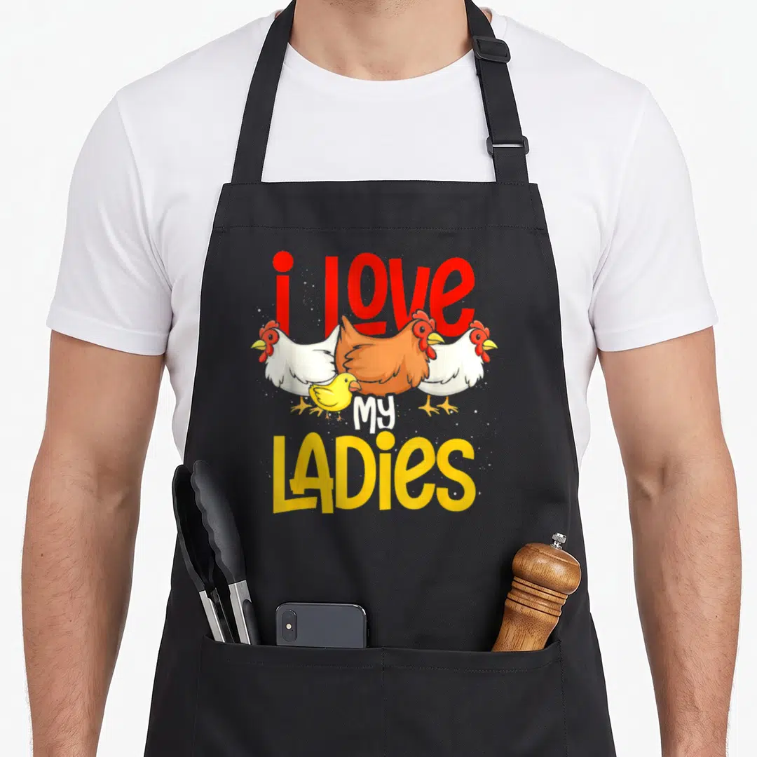 Farmer animal chicken apron - Three cartoon chickens and a chick surrounded by i love my ladies text in red and yellow — shown on a black cotton canvas apron against a white background