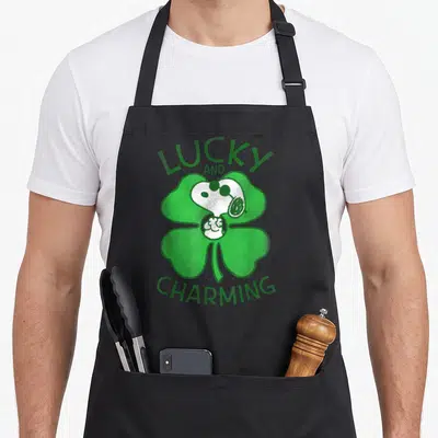 Patrick st apron - Snoopy head inside green shamrock with lucky and charming text — shown on a black cotton canvas apron against a white background