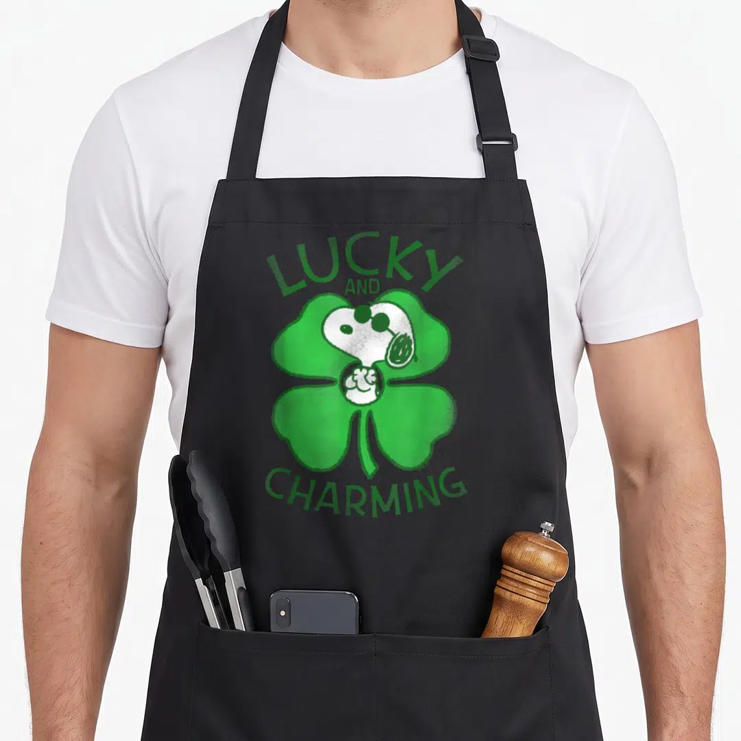 Patrick st apron - Snoopy head inside green shamrock with lucky and charming text — shown on a black cotton canvas apron against a white background