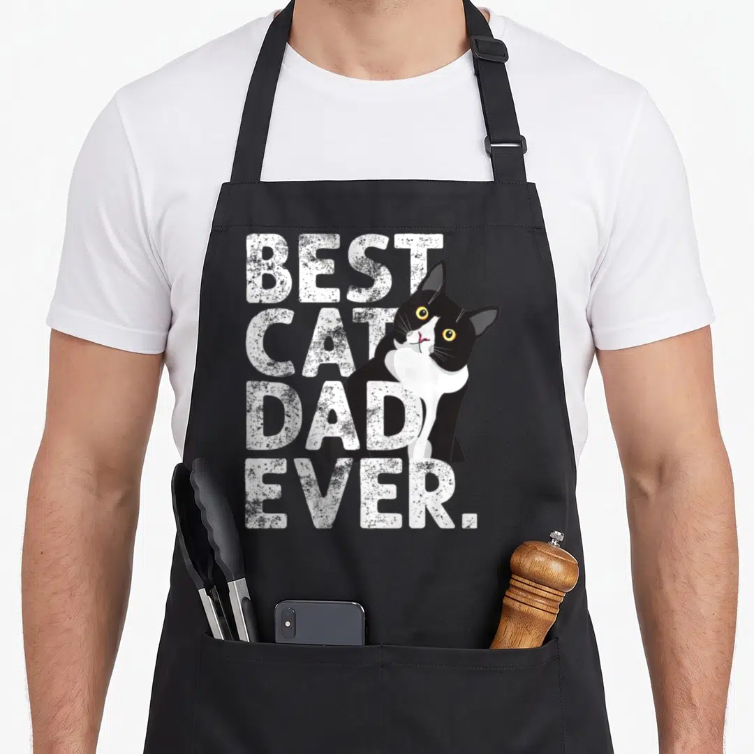 Daddy tuxedo apron - Best Cat Dad Ever text with cartoon black and white cat and yellow eyes — shown on a black cotton canvas apron against a white background