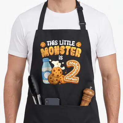 2nd cookies apron - Cartoon milk bottle, cookie cluster, and cookie-shaped number 2 with text — shown on a black cotton canvas apron against a white background