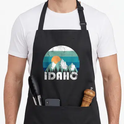 State idaho apron - Retro circular mountain landscape with orange sun and white IDAHO text on black fabric — shown on a black cotton canvas apron against a white background