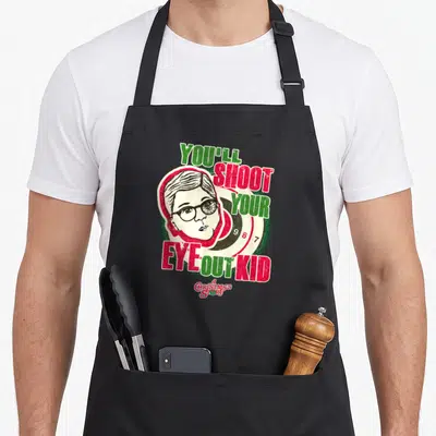 Youll christmas apron - Boy face on target with YOU'LL SHOOT YOUR EYE OUT KID text in red and green — shown on a black cotton canvas apron against a white background